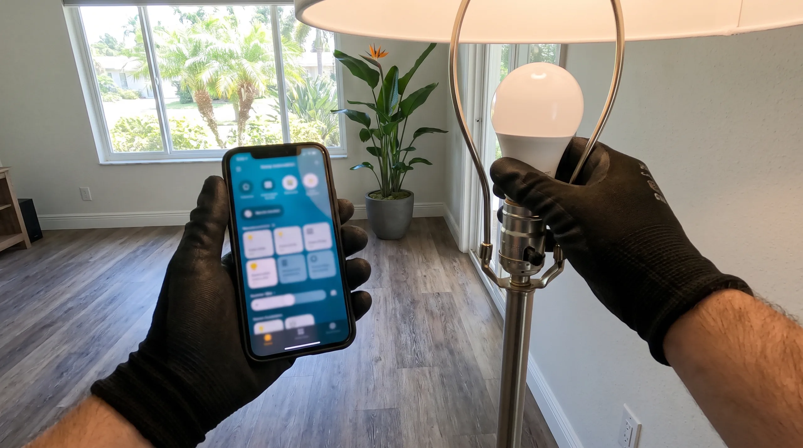 Photorealistic POV shot of clean black gloved hands holding a modern smartphone displaying a home automation app interface. The other hand is screwing a smart LED bulb into a sleek modern floor lamp base inside a clean suburban living room in a Florida home with luxury vinyl plank flooring and tropical plant decor visible through a large clean window. Strictly no faces, screen shows no legible text, bodies cropped out.