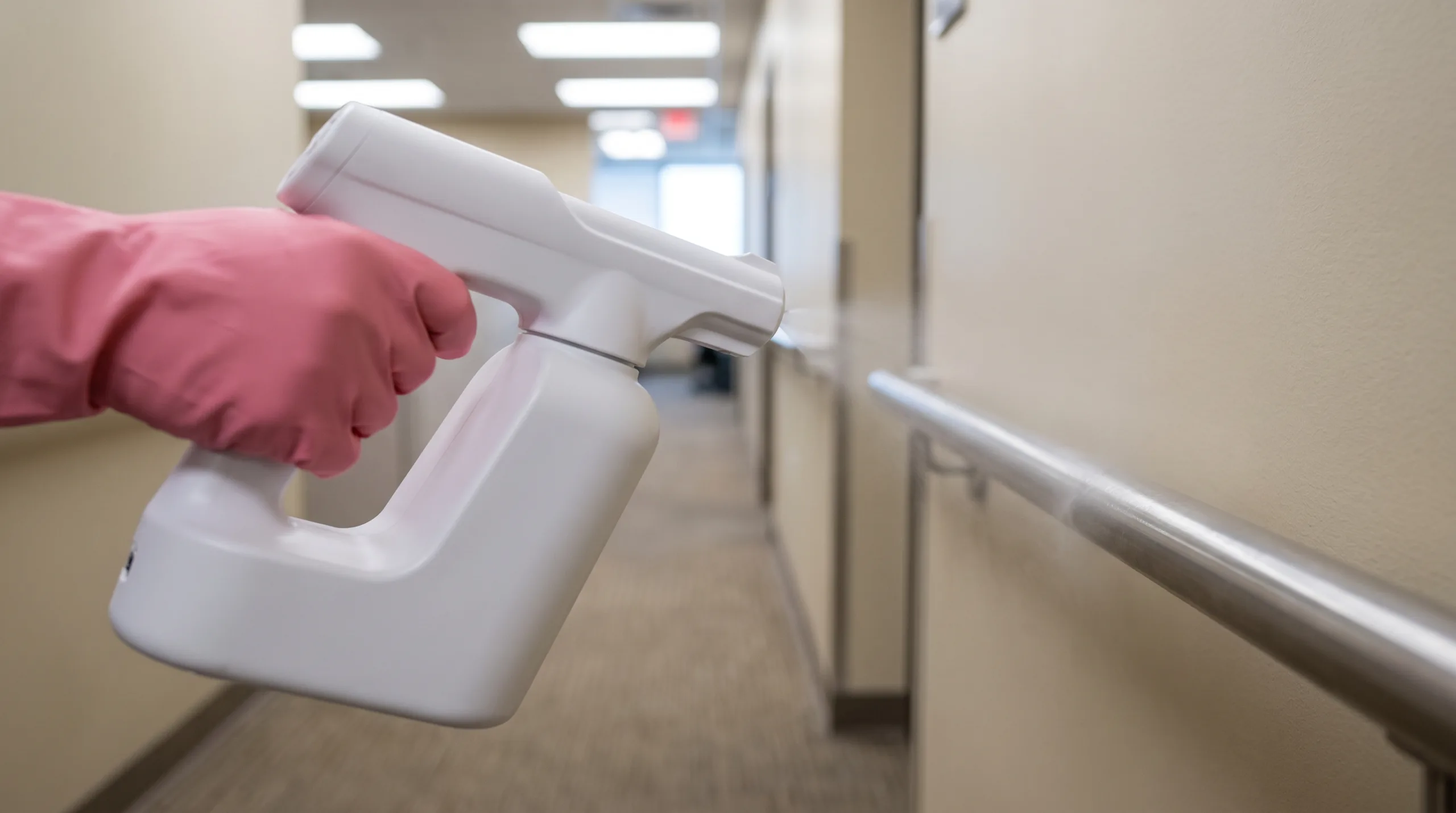Photorealistic close-up POV shot of a woman's hand wearing a pink rubber glove holding a plain, unbranded handheld electrostatic disinfection sprayer. She is spraying a fine, clean sanitizing mist directly onto a standard metal handrail in a brightly lit, typical American office hallway. The blurred background shows a clean, standard commercial space. Emphasizes hygiene and public safety on high-touch surfaces. Plain solid-color equipment, zero text, no logos. Strictly no faces, body cropped out, no plants, no text anywhere.