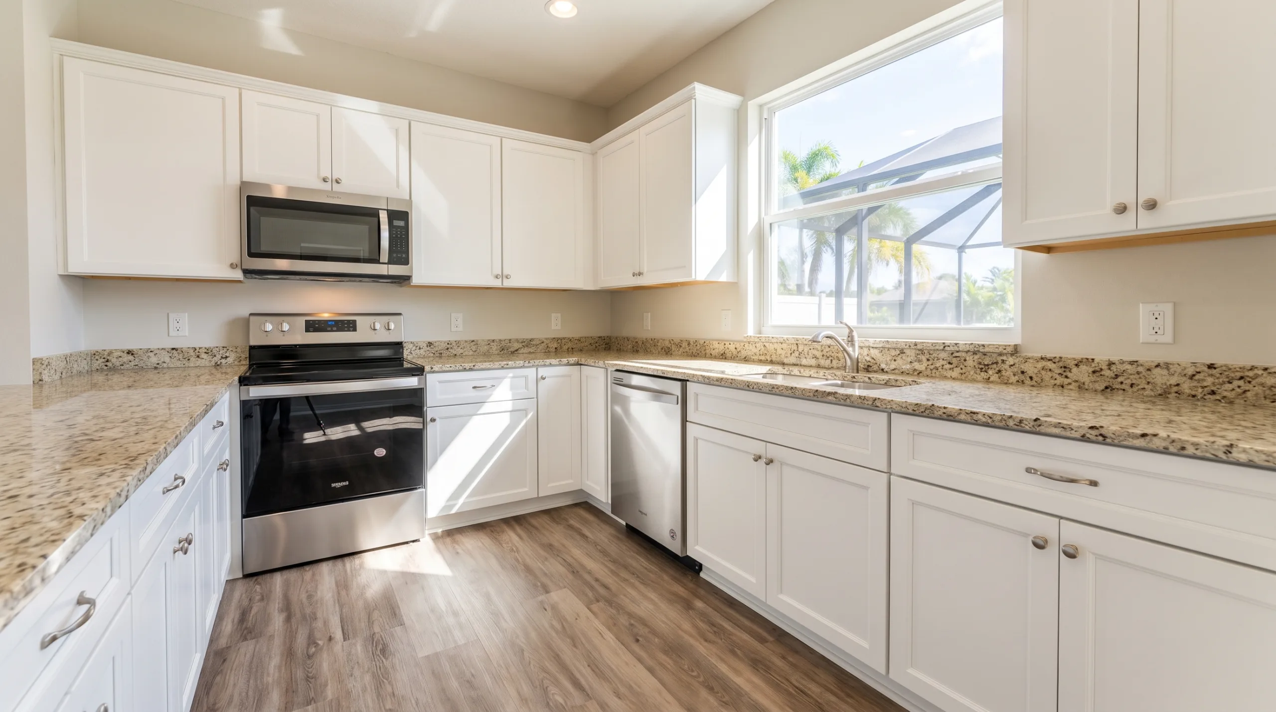 A sparkling clean, empty kitchen in a typical middle-class Florida suburban home. Features standard unbranded stainless steel appliances, simple light-colored granite countertops, and standard white wooden cabinets. Clean wood-look luxury vinyl plank flooring. Sunlit from a nearby window showing a bright day outside. Spotless surfaces, completely empty of clutter and food.