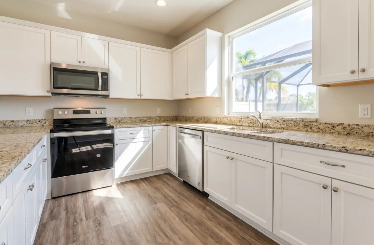 A sparkling clean, empty kitchen in a typical middle-class Florida suburban home. Features standard unbranded stainless steel appliances, simple light-colored granite countertops, and standard white wooden cabinets. Clean wood-look luxury vinyl plank flooring. Sunlit from a nearby window showing a bright day outside. Spotless surfaces, completely empty of clutter and food.